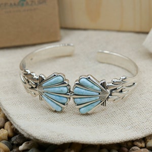May include: A silver bracelet with two blue turquoise shell designs. The bracelet is on a white fabric surface.