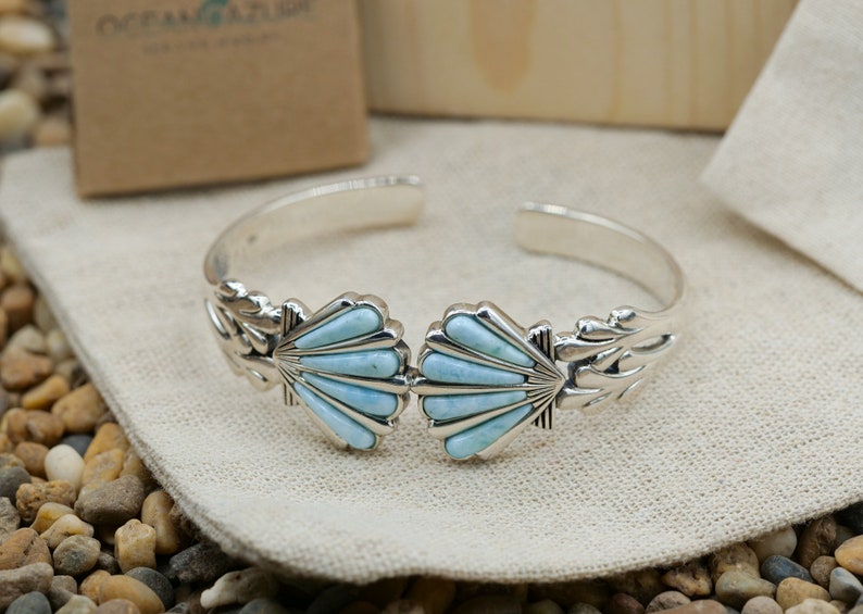 May include: A silver bracelet with two blue turquoise shell designs. The bracelet is on a white fabric surface.
