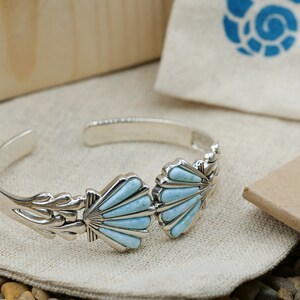 May include: A silver bracelet with two blue turquoise shell-shaped charms. The bracelet is on a white fabric background with small stones.