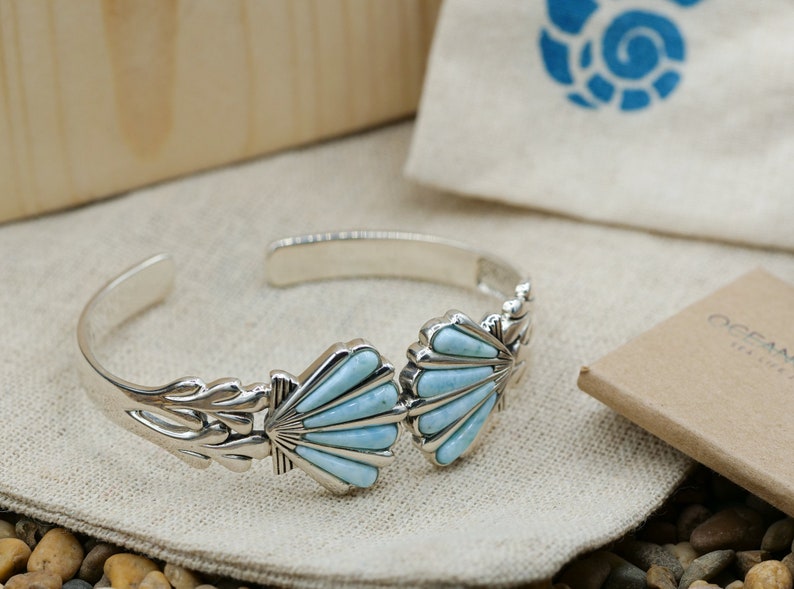 May include: A silver bracelet with two blue turquoise shell-shaped charms. The bracelet is on a white fabric background with small stones.
