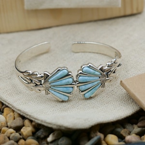 May include: A silver bracelet with a blue gemstone design. The bracelet features two shell-shaped designs with a blue gemstone inlay. The bracelet is on a light brown fabric background.