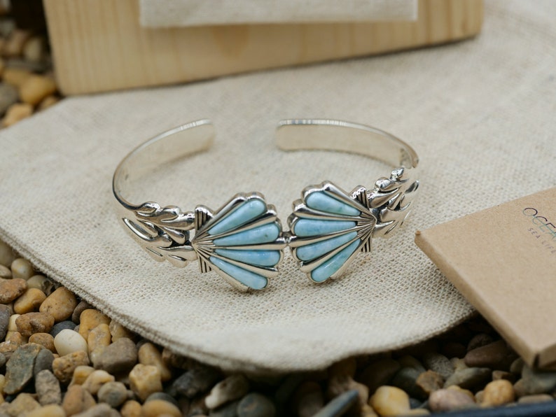 May include: A silver bracelet with a blue gemstone design. The bracelet features two shell-shaped designs with a blue gemstone inlay. The bracelet is on a light brown fabric background.