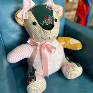 May include: A patchwork teddy bear with pink and white striped fabric, green floral fabric, and yellow fabric. The bear has pink bows on its ears and a pink ribbon around its neck. The bear is sitting on a blue armchair. The bear's foot is embroidered with the name "Lottie".