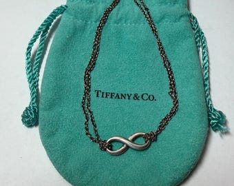 Puede incluir: Una pulsera de color plateado con el símbolo de infinito y una cadena doble fina, sobre una bolsa de tela suave de color verde azulado con cordón, que presenta el nombre de la marca Tiffany & Co. impreso en letras negras al frente.