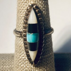 May include: A silver ring with a turquoise, black, and white stone inlay. The stone is set in a silver bezel and surrounded by a row of silver beads.