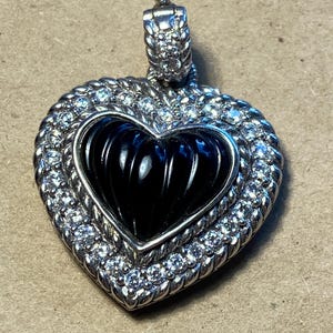 Puede incluir: Colgante en forma de coraz&oacute;n con centro negro y marco plateado. El marco est&aacute; incrustado con peque&ntilde;as piedras transparentes. La fianza tambi&eacute;n es plateada y tiene un dise&ntilde;o texturizado. El colgante probablemente est&aacute; hecho de metal y piedras preciosas.