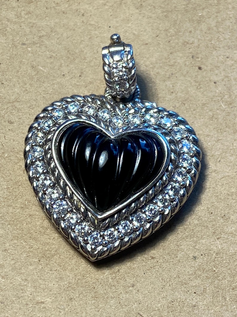 Puede incluir: Colgante en forma de coraz&oacute;n con centro negro y marco plateado. El marco est&aacute; incrustado con peque&ntilde;as piedras transparentes. La fianza tambi&eacute;n es plateada y tiene un dise&ntilde;o texturizado. El colgante probablemente est&aacute; hecho de metal y piedras preciosas.