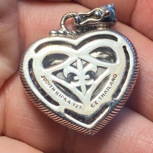 Puede incluir: Colgante plateado en forma de coraz&oacute;n con dise&ntilde;o de flor de lis. El colgante tiene un borde texturizado y el texto "JUDITH RIPKA 925 CZ THAILAND" es visible. La fianza es una cabeza de drag&oacute;n.