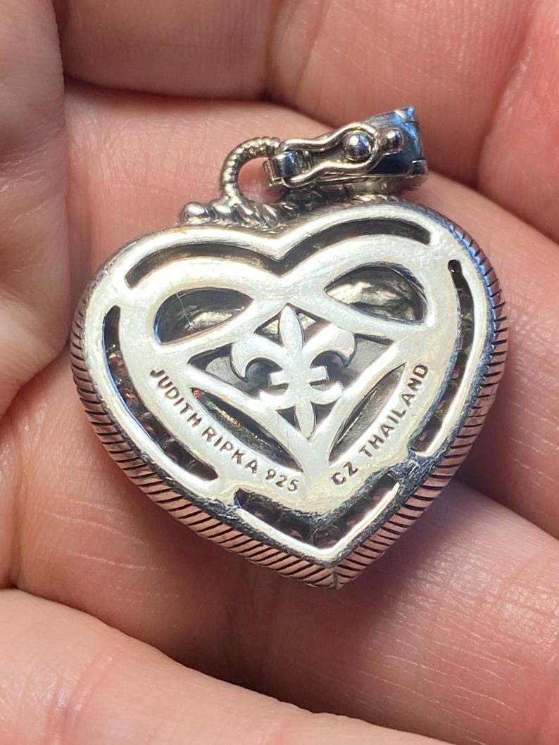 Puede incluir: Colgante plateado en forma de coraz&oacute;n con dise&ntilde;o de flor de lis. El colgante tiene un borde texturizado y el texto "JUDITH RIPKA 925 CZ THAILAND" es visible. La fianza es una cabeza de drag&oacute;n.