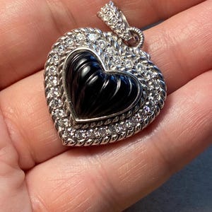 Puede incluir: Colgante en forma de coraz&oacute;n con un centro negro y un borde de peque&ntilde;as piedras preciosas transparentes. El colgante es plateado y tiene un borde texturizado en forma de cuerda. La fianza tambi&eacute;n es plateada y est&aacute; adornada con peque&ntilde;as piedras preciosas.