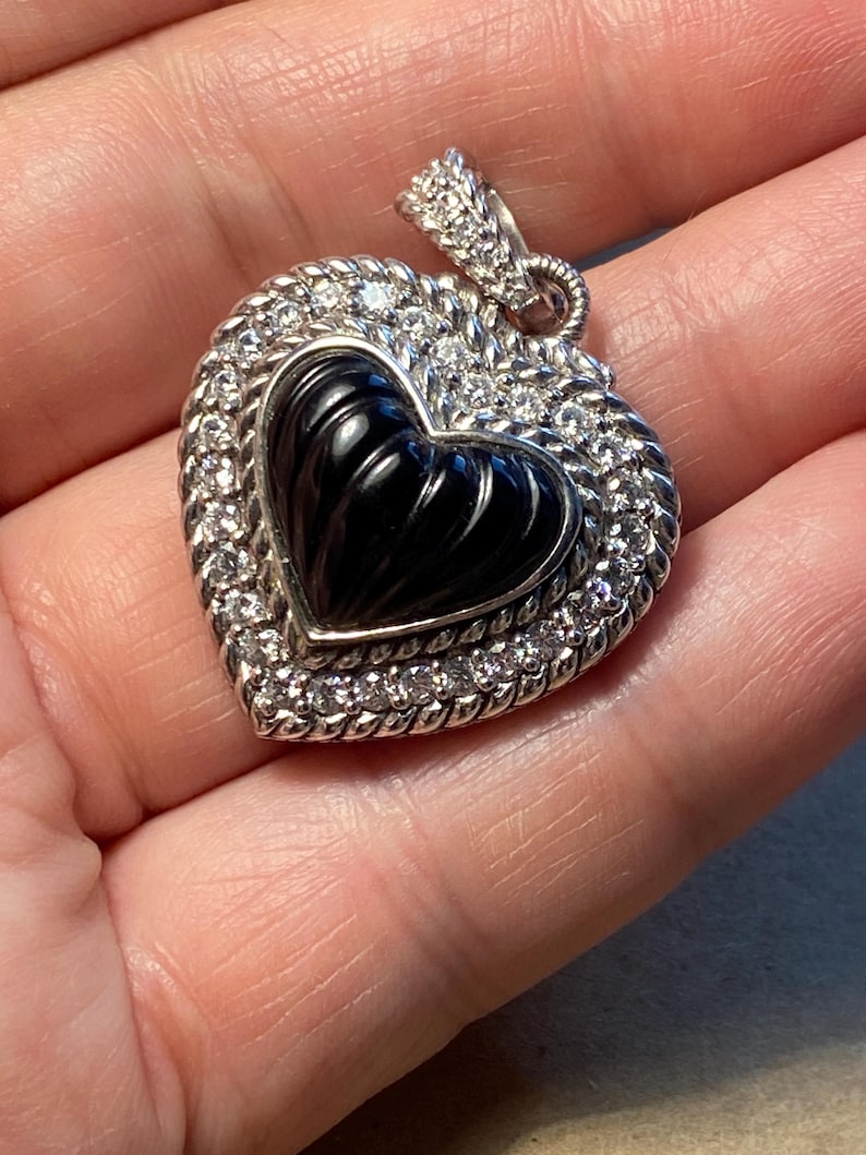 Puede incluir: Colgante en forma de coraz&oacute;n con un centro negro y un borde de peque&ntilde;as piedras preciosas transparentes. El colgante es plateado y tiene un borde texturizado en forma de cuerda. La fianza tambi&eacute;n es plateada y est&aacute; adornada con peque&ntilde;as piedras preciosas.