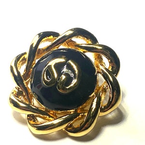 May include: A gold-toned brooch with a central black enamel circle featuring a gold design. The brooch has a woven, floral-like design around the center. The jewelry piece is set against a white background.