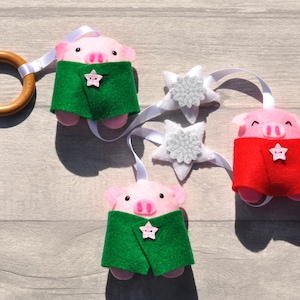 Puede incluir: Móvil para bebé hecho a mano con tres cerdos rosas de fieltro que llevan capas verdes y rojas, adornados con botones en forma de estrella. Estrellas de fieltro blanco con centros de copos de nieve plateados y un anillo de madera.