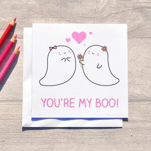 May include: A white card with two cartoon ghosts in pink and white. The ghost on the left is holding its hands together and the ghost on the right is holding a pink flower. The text "YOU'RE MY BOO!" is written in pink letters at the bottom of the card.