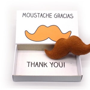 Puede incluir: Una caja blanca con un bigote de fieltro marrón en su interior. La caja tiene las palabras "Moustache Gracias" y "Thank You!" impresas en ella. La caja está abierta y el bigote es visible.