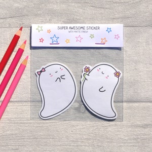 May include: Two white ghost stickers with cute faces. One ghost is wearing a pink bow and the other is holding a pink flower. The stickers are on a white background with a header that says "Super Awesome Sticker with Matte Finish".