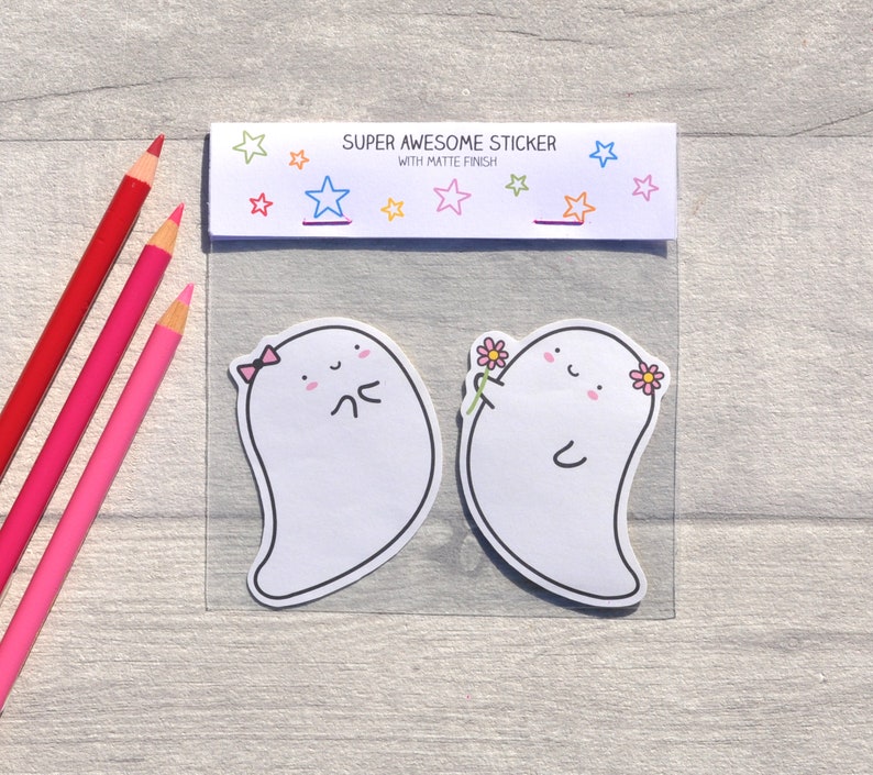 May include: Two white ghost stickers with cute faces. One ghost is wearing a pink bow and the other is holding a pink flower. The stickers are on a white background with a header that says "Super Awesome Sticker with Matte Finish".