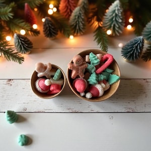 May include: Two wooden bowls filled with Christmas-themed wax melts. The melts include gingerbread men, Santa hats, candy canes, and Christmas trees in shades of pink, green, and brown. The bowls sit on a white wooden surface with Christmas tree branches and lights in the background.