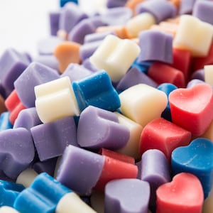 May include: A selection of heart-shaped wax melts in blue, purple, pink, white and beige colours. The wax melts are arranged in a pile on a white surface.