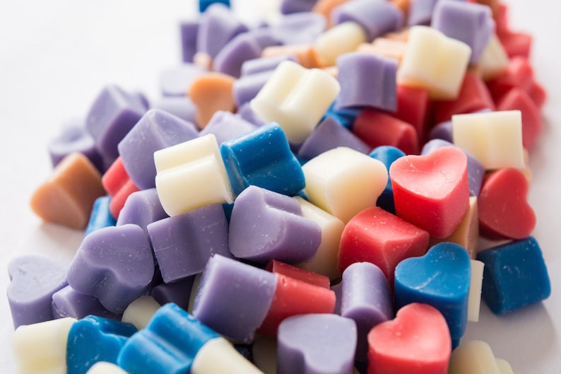 Puede incluir: Una variedad de fundentes de cera en forma de coraz&oacute;n en colores azul, morado, rosa, blanco y beige. Los fundentes de cera est&aacute;n dispuestos en una pila sobre una superficie blanca.