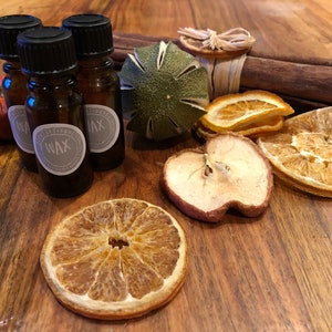 May include: Three small brown glass bottles with black lids and labels that say "Essential Wax" on a wooden surface with dried orange slices, a dried apple slice, a green citrus fruit, and cinnamon sticks.