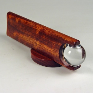 May include: A wooden kaleidoscope with a clear glass sphere at one end. The kaleidoscope is made of dark red wood with a natural wood grain pattern. The sphere is mounted on a circular base.