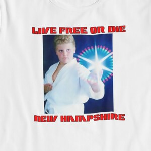 Puede incluir: Camiseta blanca con un gráfico rojo de una persona joven en una pose de karate con el texto "Live Free or Die" y "New Hampshire" debajo de la imagen.