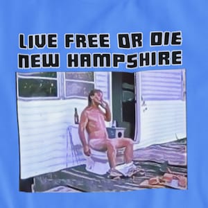 Puede incluir: Una camiseta azul claro con el texto "Live Free or Die New Hampshire" impreso en la parte delantera. La camiseta presenta una foto de un hombre sentado en un inodoro fuera de una caravana.