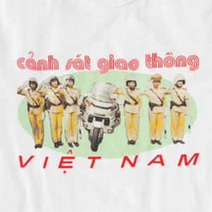 May include: White t-shirt with a vintage-style graphic of Vietnamese traffic police officers saluting, riding a motorcycle, and the text "cảnh sát giao thông" and "VIETNAM" in red.