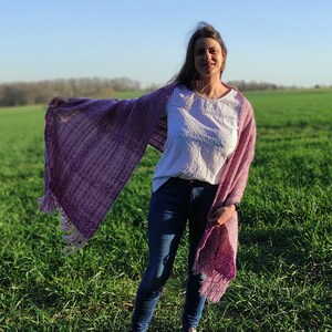 Könnte beinhalten: Eine Frau in einem weißen Shirt und blauen Jeans steht auf einem Feld mit grünem Gras. Sie trägt einen lila-weißen Schal mit Fransen. Der Schal ist über ihre Schultern gelegt und weht im Wind.