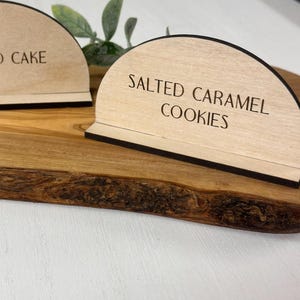 May include: Two semi-circular light wood signs with black edges. The left sign features the black text ALMOND CAKE, and the right sign features the black text SALTED CARAMEL COOKIES. Both signs are positioned on a wooden board with a natural edge.