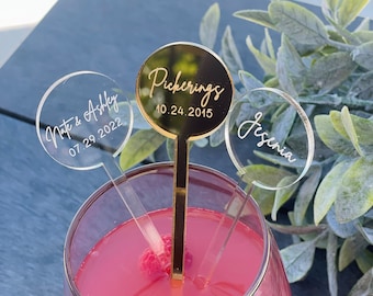 May include: Three clear acrylic drink stirrers with gold lettering. The stirrers have the names "Nate & Ashley 07.29.2022", "Pickering's 10.24.2015", and "Jesenia" on them. The stirrers are in a glass with a pink drink and raspberries.
