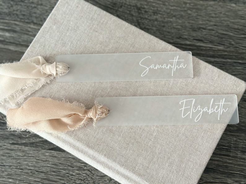 May include: Two clear acrylic bookmarks with the names "Samantha" and "Elizabeth" written in white script. Each bookmark is tied with a beige ribbon.
