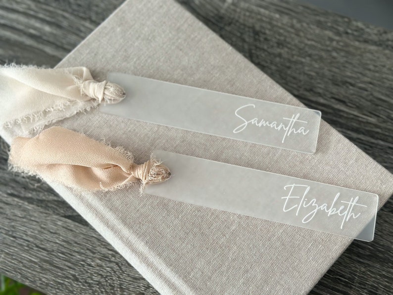 May include: Two clear acrylic bookmarks with the names "Samantha" and "Elizabeth" written in white cursive. Each bookmark is tied with a beige ribbon.