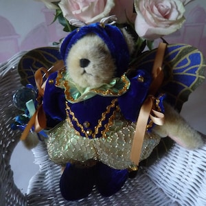 May include: A plush teddy bear dressed in a blue velvet outfit with gold trim and a matching hat. The bear has large, ornate blue and gold wings. A bouquet of pink roses sits behind the bear, which is in a white wicker basket.
