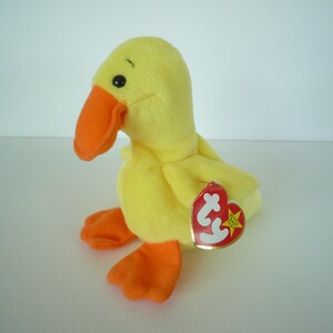 May include: A yellow plush duck toy with an orange beak and feet. The duck has a red heart-shaped tag attached to its side. The toy is made of soft material and is designed to resemble a duck.