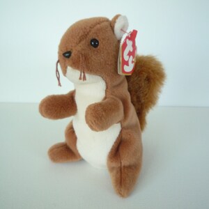 May include: A brown plush squirrel toy with a white belly and a fluffy tail. The squirrel has small black eyes, whiskers, and a red tag attached to its ear. The toy is sitting on a white surface.