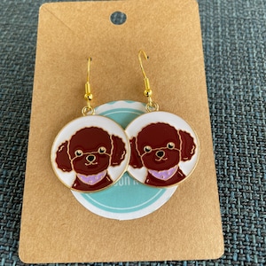 May include: A pair of gold dangle earrings with white circular charms featuring a brown poodle with a purple collar. The earrings are on a brown card with the text "Sun F" on it.