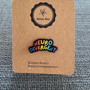 Puede incluir: Un pin de esmalte negro con las palabras "Neuro Divergente" en colores arcoíris. El pin está en una tarjeta marrón con el texto "Addison Rose Co" y "addisonrosejewelleryco" en negro.