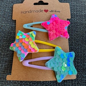 Peut inclure: Ensemble de trois pinces à cheveux en forme d'étoile avec des paillettes colorées. Les pinces sont sur une carte brune avec le texte "Handmade with love".