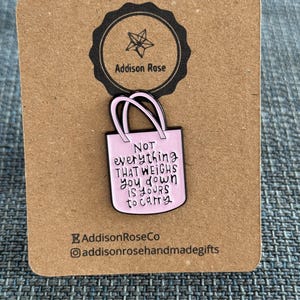 Op de afbeelding: Een roze emaille pin in de vorm van een draagtas met de tekst "Not everything that weighs you down is yours to carry". De pin is bevestigd op een bruine kaart met het Addison Rose logo.