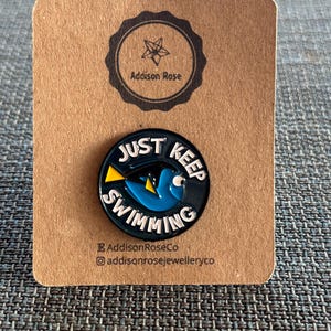 May include: A round, black enamel pin with the words "JUST KEEP SWIMMING" encircling a blue fish with yellow fins. The pin is on a brown card with the text "Addison Rose" and social media handles.