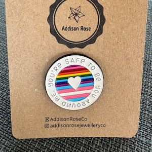 Op de afbeelding: Een ronde, witte emaille pin met de tekst "YOU'RE SAFE TO BE YOU AROUND ME". De pin heeft een regenboogstreepontwerp met een wit hart in het midden. De pin zit op een bruine kaart met de tekst "Addison Rose".