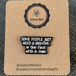 May include: A black enamel pin with white text that reads "SOME PEOPLE JUST NEED A HIGH-FIVE in the face. with a chair." The pin is on a brown card with the Addison Rose logo and social media handles.