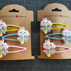 Puede incluir: Dos juegos de pinzas para el pelo con un diseño de nube arcoíris. Cada juego incluye tres pinzas con bases de diferentes colores. El embalaje es de cartón marrón con el texto "Handmade with love".