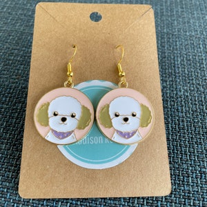 May include: A pair of gold-tone earrings featuring a white poodle with pink and green accents. The earrings are on a brown card with the text "Madison K" in white.