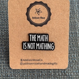 May include: A black and white enamel pin with the text "THE MATH IS NOT MATHING" on a brown card. The card has a circular logo with a star and the words "Addison Rose". The bottom of the card has the text "AddisonRoseCo" and an Instagram handle.