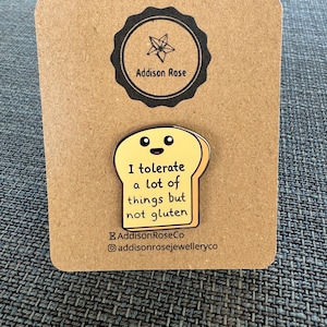 May include: A yellow enamel pin shaped like a slice of bread with a smiling face. The pin says "I tolerate a lot of things but not gluten". The pin is on a brown card with a black and white logo that says "Addison Rose".