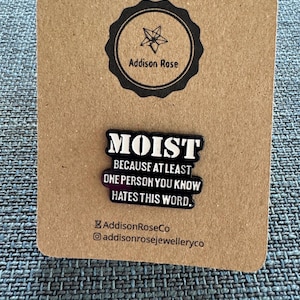 May include: Black enamel pin with white text that reads "MOIST BECAUSE AT LEAST ONE PERSON YOU KNOW HATES THIS WORD." The pin is on a brown card with the text "Addison Rose" in a circle.