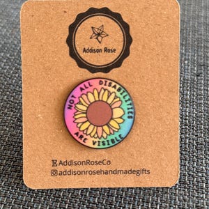 May include: A circular pin featuring a sunflower design and the words "NOT ALL DISABILITIES ARE VISIBLE". The pin is on a brown card with the "Addison Rose" logo and social media details.